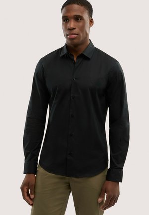 Mężczyzna ubrany w dopasowaną czarną koszulę na guziki z długimi rękawami i brązowe spodnie, stojący na jednolitym jasnym tle.