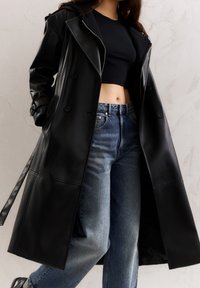 Cazadora larga de imitación piel negra con botones de doble botonadura, solapas anchas y una textura suave, combinada con unos pantalones vaqueros de cintura alta en azul claro.