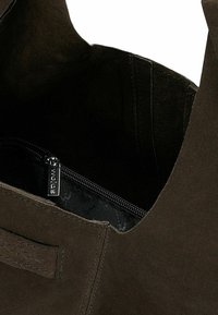 Borsa in camoscio marrone con un'ampia apertura, dotata di una tasca interna con cerniera. L'interno presenta una fodera nera e dettagli con il logo.