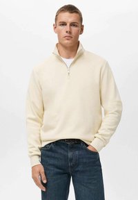Cream-colored zip-up sweatshirt with a stand collar, made of soft material, paired with dark blue jeans. Smooth texture with no patterns.