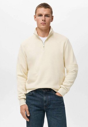 Sudadera cremosa con cremallera y cuello alto, hecha de material suave, combinada con jeans de color azul oscuro. Textura lisa sin patrones.