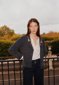 Dunkelgrauer Cardigan mit floraler Stickerei auf den Schultern, weißes Blusenshirt, hoch taillierte schwarze Hose und geschichtete Halsketten, vor einem grünen Hintergrund.