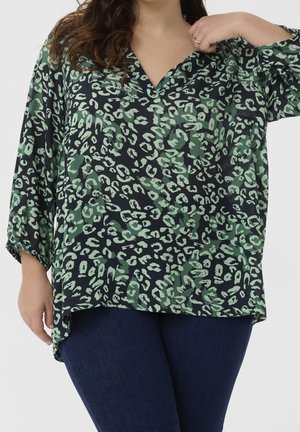 Vrouw draagt een donkergroene en zwarte dierenprintblouse met driekwart mouwen en donkerblauwe spijkerbroek tegen een effen achtergrond.