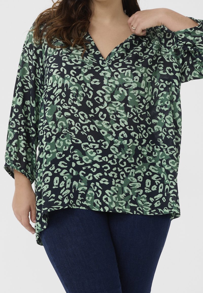 Femme portant un chemisier imprimé animal vert foncé et noir à manches trois-quarts et un jean bleu foncé sur un fond uni.