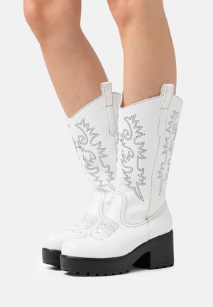 Valkoiset cowboy-saappaat, joissa on mustaa kirjailua, pyöreä kärki ja paksu musta korko. Valmistettu sileästä materiaalista, jossa on kohokuvioitua ompelua.