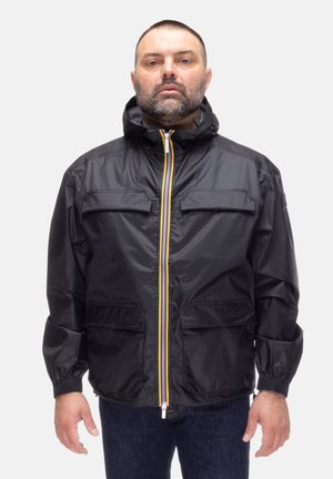 Uomo con barba corta che indossa una giacca nera con cappuccio, cerniera con strisce gialle e polsini elasticizzati, in piedi davanti a uno sfondo uniforme.