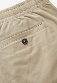 A khaki nadrág rugalmas derékkal, egy hátsó zseb fekete gombbal és sima, textúrált anyaggal rendelkezik.