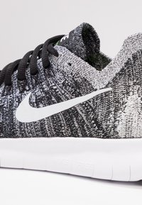 Sneaker in maglia bianca e nera con una parte superiore texturizzata, caratterizzata da un logo Nike bianco e una suola flessibile bianca con branding visibile.