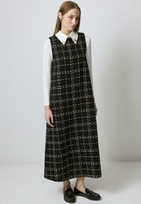 Žena u crnoj bezrukavnoj kariranoj dugoj haljini preko bijele dugosleeve košulje s velikim ovratnikom i crnim mokasinkama, stoji.
