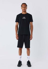 Ensemble t-shirt noir et shorts, en tissu doux. Le t-shirt présente un logo blanc central ; les shorts sont à hauteur des genoux avec des détails sur les côtés. Sneakers noires et chaussettes blanches.