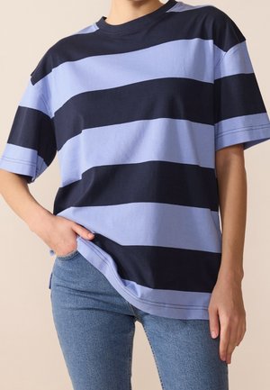 Persoon draagt een losse T-shirt met korte mouwen met dikke horizontale donkerblauwe en lichtblauwe strepen, gecombineerd met een blauwe spijkerbroek.