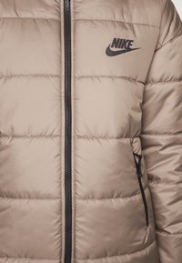 Tan puffer jacket with quilted design, featuring a black zipper and side pocket. Black Nike logo on chest. Smooth, shiny fabric finish.