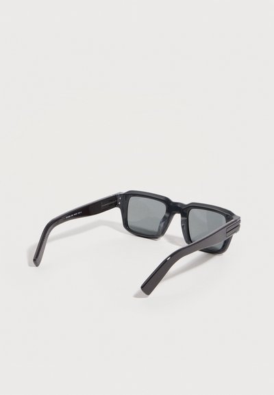 Lunettes de soleil rectangulaires noires avec des montures épaisses et des verres foncés reposant sur une surface claire, branches légèrement ouvertes.