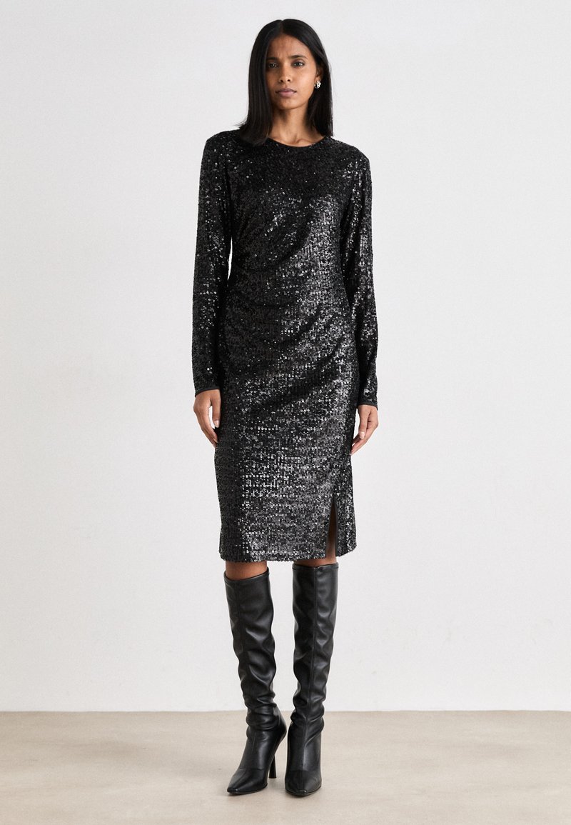 Vestido preto com lantejoulas, mangas longas e uma fenda lateral. O material é brilhante com uma superfície texturizada. Combinado com botas altas pretas até ao joelho.