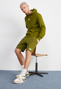 Completo con felpa verde oliva e pantaloni corti, con logo North Face; abbinato a sneakers bianche e beige. Tessuto morbido, vestibilità comoda, design casual.