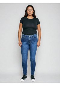 Pikkade tumejuustega naine seisab näoga otse kaamera poole, kandes musta T-särki, siniseid kõrge vöökohaga kitsaid teksapükse ja musti tenniseid, ilma efekti või taustata.