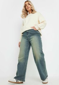 Kermaru sweatshirtilla, jossa on roikkuvat olkapäät, yhdistettynä haalistuneisiin vihreisiin leveälahkeisiin farkkuihin, joissa on korkea vyötärö ja hienovarainen väri-gradientti. Valkoiset lenkkarit.