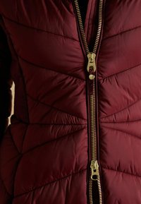 Burgundi värvi tikanditega puffjakker, millel on kuldne tõmblukk, diagonaalsed õmblused ja sile, läikiv tekstuur.