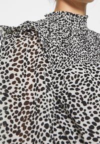 Vit blus med ett svart abstrakt prickigt mönster, med en rynkad hög halsringning och smockad midja. Genomskinligt, lätt tyg med flödande  ärmar.