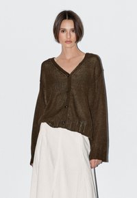 Jonge vrouw met kort bruin haar, gekleed in een losgebreid bruin vest over een witte rok met hoge taille, staand tegen een effen witte achtergrond.