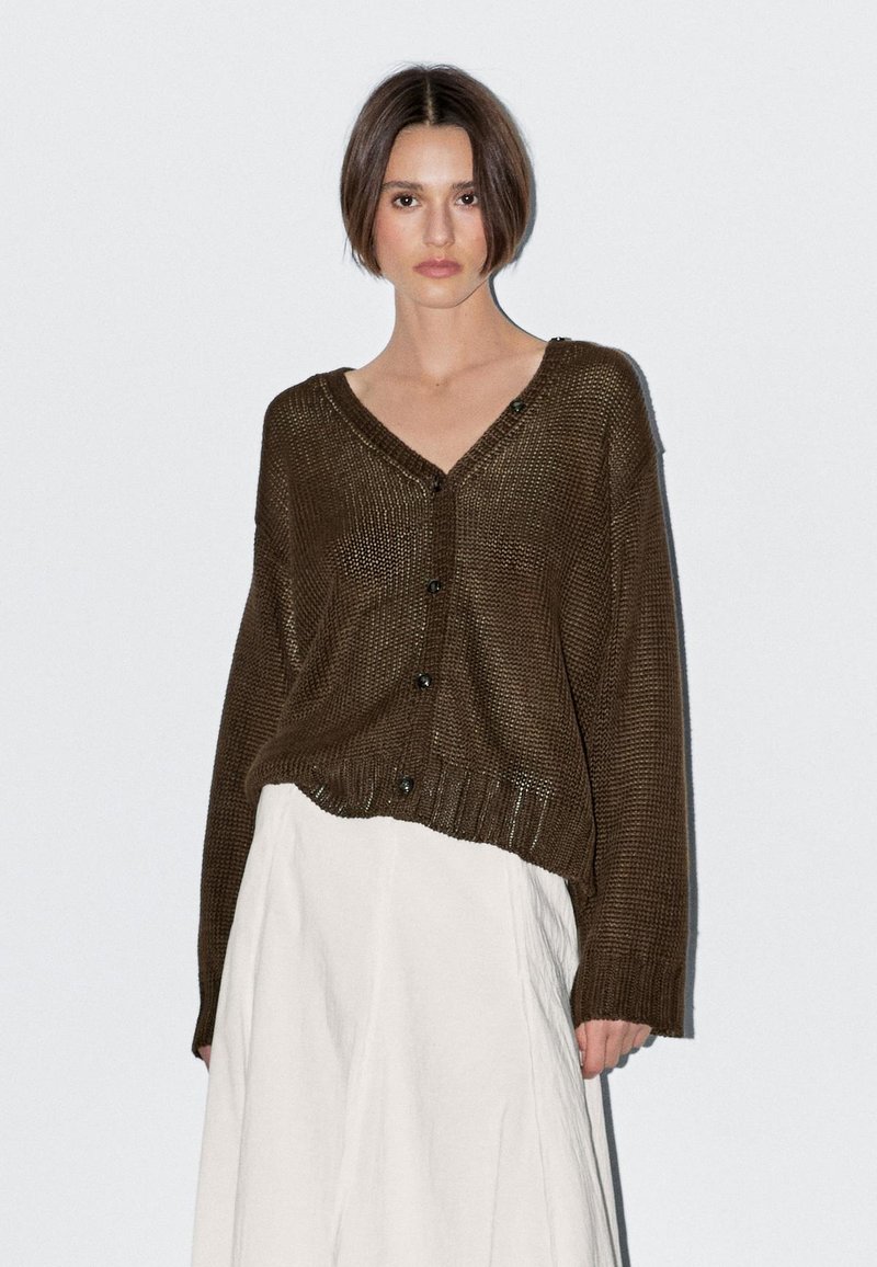 Jonge vrouw met kort bruin haar, gekleed in een losgebreid bruin vest over een witte rok met hoge taille, staand tegen een effen witte achtergrond.