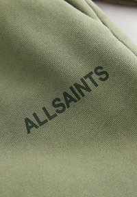Zielona bawełniana tkanina o teksturowanej powierzchni, z nadrukowaną nazwą marki "ALLSAINTS" w ciemnozielonym kolorze. Prosty i minimalistyczny design.