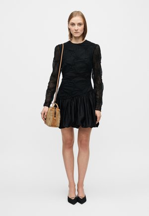 Μαύρο φόρεμα με υφή υφάσματος, μακριά διαφανή μανίκια και μια φούστα με πιέτες που ανοίγει προς τα έξω. Το μοντέλο κρατάει μια μικρή τσάντα σε καφέ και μπεζ αποχρώσεις με λουράκι.