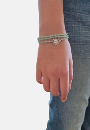 Polso con braccialetto a tre fili di perline verdi con un ciondolo rotondo in argento con un motivo geometrico, su sfondo di jeans blu.