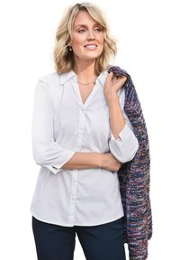 Camicia bianca con maniche a tre quarti, scialle texturizzato viola e blu drappeggiato su una spalla, indossato sopra pantaloni scuri.