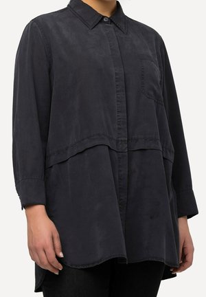 Chemise grise foncée à manches longues avec poche frontale, col boutonné et ourlet à couches superposées portée par une personne avec mains et torse visibles.