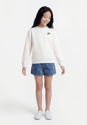 Tüdruk, kes kannab valget Nike fliisipaela, teksapükse väikese lilleornamendiga ja valgeid tossusid, seistes ja allapoole vaatades naeratades.