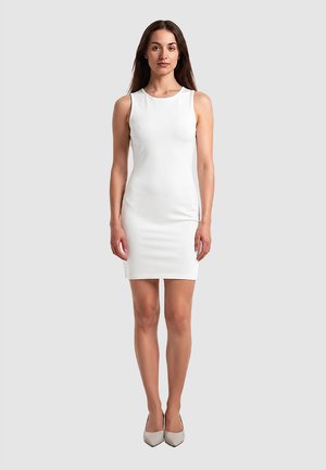 Donna in piedi, rivolta in avanti, indossa un vestito bianco senza maniche al ginocchio e tacchi chiari, su uno sfondo grigio semplice.