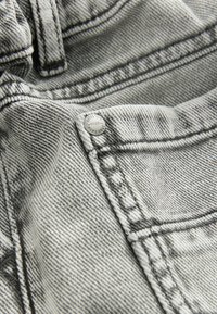 Gros plan sur un tissu en denim gris clair avec des coutures visibles, un rivet métallique embossé de "AUTHENTIC 1982 NEXT DOOR" et un détail de poche.