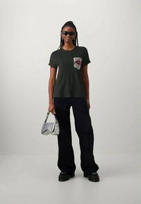 Σκούρο πράσινο T-shirt με παγιέτα στην τσέπη, που απεικονίζει ένα σχέδιο με κόκκινα χείλη. Συνδυάζεται με χαλαρές μαύρες παντελόνες και ασημένια τσάντα.
