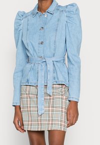 Veste en denim bleu clair avec épaules bouffantes, fermeture à boutons et taille ceinturée. Associée à une mini-jupe à carreaux dans des tons chauds.