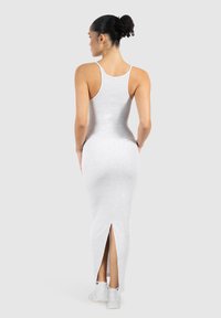 Kvinde med krøllet knold iført en tætsiddende lysegrå ærmeløs maxi-kjole med slids bagpå og hvide sneakers, stående med ryggen til.