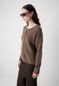Cardigan marron texturé avec un col rond, fermeture par boutons et manches longues, associé à une jupe marron côtelée avec une fente sur le côté.