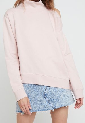 Nainen, joka käyttää vaaleanpunaista kylmillä olkapäillä varustettua collegepaitaa korkealla kaula-aukolla ja vaaleansinistä happopesua saanutta farkkujen minihame franssireunuksella.