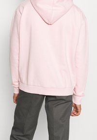 Jasnoróżowy hoodie z kieszenią kangurką, żebrowanymi mankietami i dołem, z dużym kapturem i miękkim, teksturowanym materiałem. Noszony z szarymi spodniami.