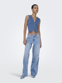 Jonge vrouw staat met ontspannen handen, draagt een mouwloos denimvest, lichtblauwe wijd uitlopende jeans en puntige blauwe schoenen tegen een effen achtergrond.