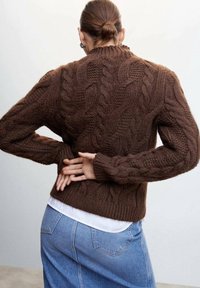 Pull en maille torsadée marron avec un col et un ourlet côtelés. Présente des motifs en points texturés et des manches longues. Porté par-dessus une chemise blanche et un jean en denim.