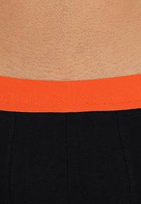 Boxer brief neri con una cintura arancione texturizzata che presenta un branding in rilievo. Inquadratura ravvicinata che mette a fuoco la cintura e la pelle.