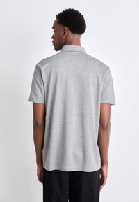 Pilkas trumpomis rankovėmis polo marškinėliai su apykakle, lygia tekstūra ir subtiliu melange raštu, dėvimi su juodomis kelnėmis, matomi iš nugaros.