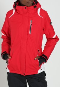 Rote wasserdichte Jacke mit weißen Akzenten, hohem Kragen, Reißverschlusstaschen und elastischen Bündchen. Aus einem strapazierfähigen, strukturierten Stoff gefertigt.