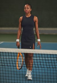 Φόρεμα τένις σε σκούρο ναυτικό ύφασμα, αμάνικο με πλαϊνές σχισμές, συνδυασμένο με λευκά βραχιόλια και παπούτσια, κρατώντας μια πορτοκαλί ρακέτα τένις.