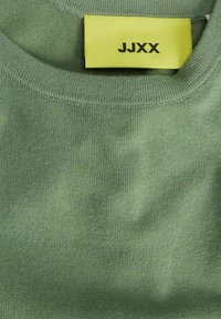 Close-up van een groene gebreid trui kraag met een geel kledinglabel met de tekst "JJXX" gestikt aan de binnenkant van de halslijn.