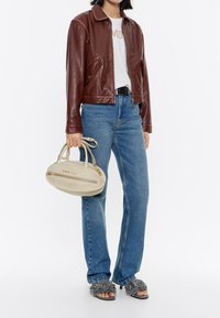 Giubbotto di pelle marrone, t-shirt bianca con testo, jeans blu, sandali grigi con texture, borsa beige con hardware dorato e logo.