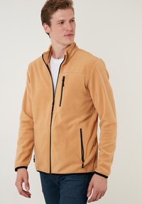 Fleece jack in lichtbruin met zwarte ritsen en accenten. Beschikt over een hoge kraag, zijzakken en een ritsvak op de borst.