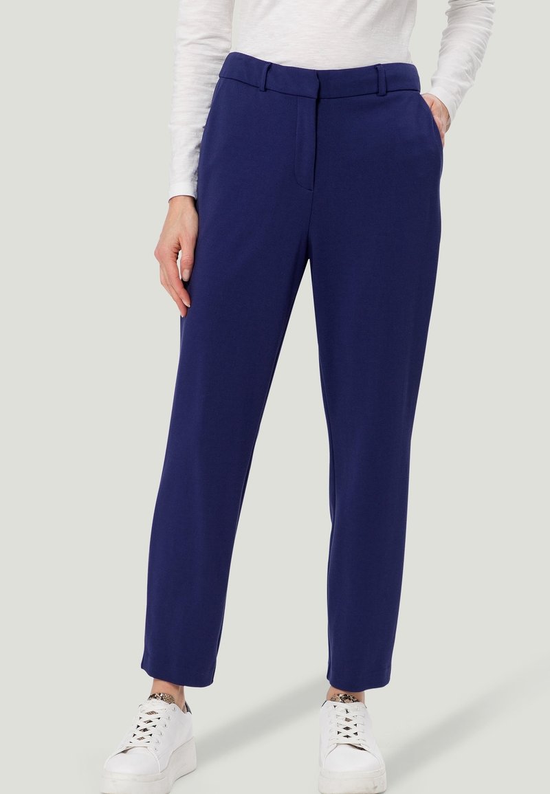 Pantalon bleu marine, ajusté, avec une texture lisse, présentant un devant plat, des poches latérales et une coupe slim, associé à des baskets blanches.