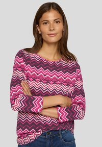 Lila und rosa Chevron-muster Shirt mit langen Ärmeln, aus strukturiertem Stoff, mit Rundhalsausschnitt und aufgerollten Ärmeln, kombiniert mit blauen Jeans.
